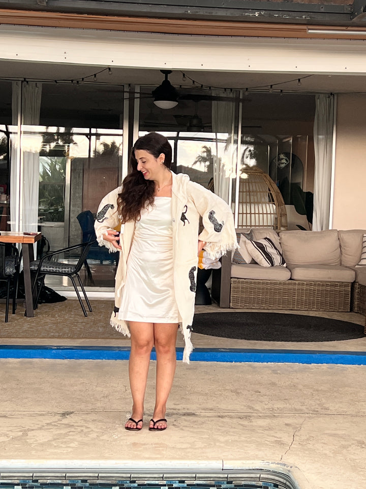 A woman in a white dress and Black Leopard Hooded Cotton Kimono Robe from turkanhome.com stands smiling near a pool, with outdoor furniture and large windows in the background.
