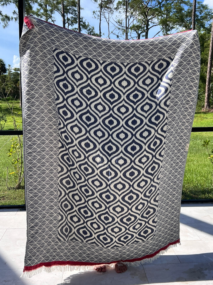 A person holds up the Ikat Throw from turkanhome.com, a large blue and white reversible geometric blanket with red corner fringe, standing indoors before windows overlooking green grass and tall trees.