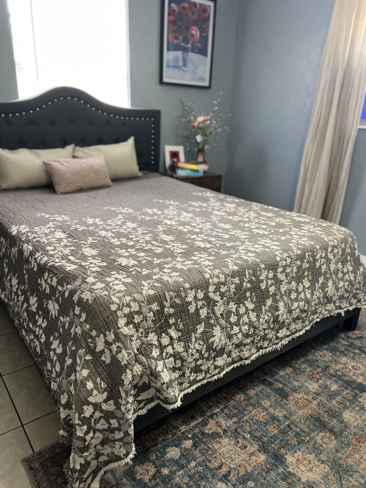 A neatly made bed features a brown Bougainvillea Muslin Bed Blanket by turkanhome.com (Queen), decorative pillows, and a dark studded headboard. A nightstand with books, flowers, and photo sits nearby, set against light blue walls with art.