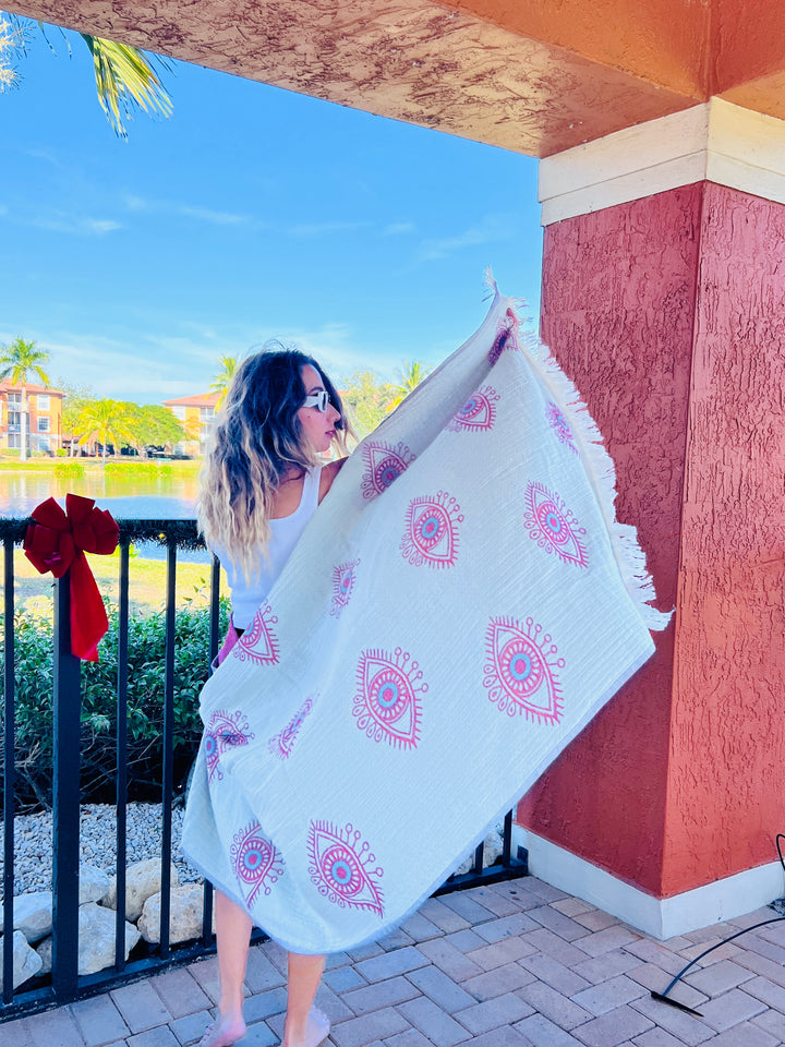 A woman with wavy hair stands barefoot on a patio, wrapped in the Eye Red Sand Resistant Turkish Beach Towel by turkanhome.com. Behind her are a black fence, lake, palm trees, and colorful buildings under a bright blue sky.