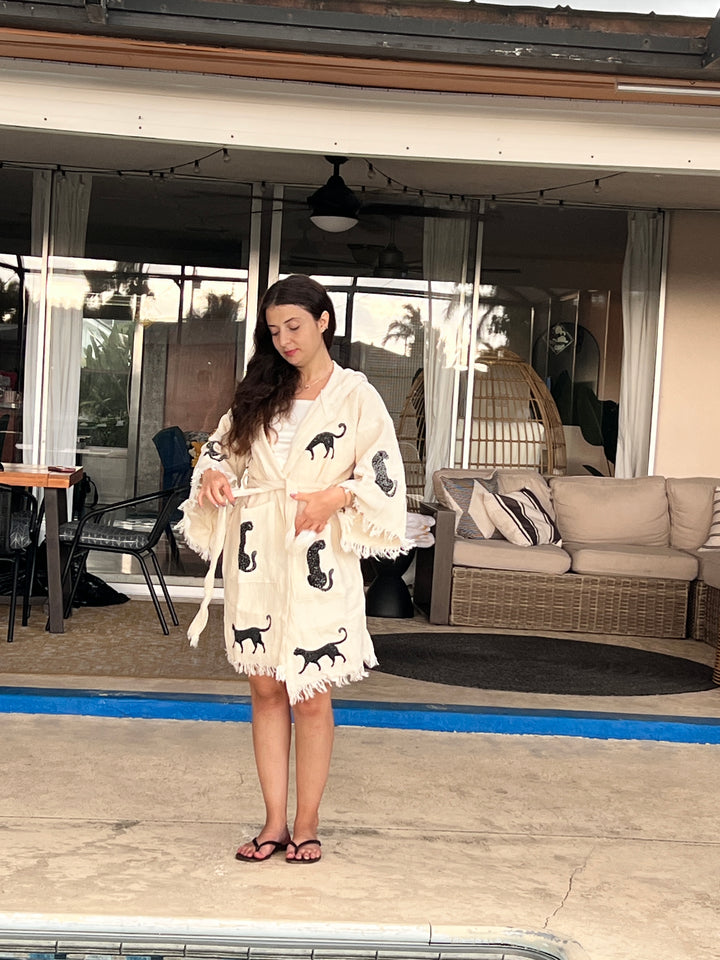 A woman with long dark hair stands by a pool wearing the Black Leopard Hooded Cotton Kimono Robe from turkanhome.com. She holds the belt and looks down, with patio chairs, a sofa, and palm trees reflected in a glass door behind her.