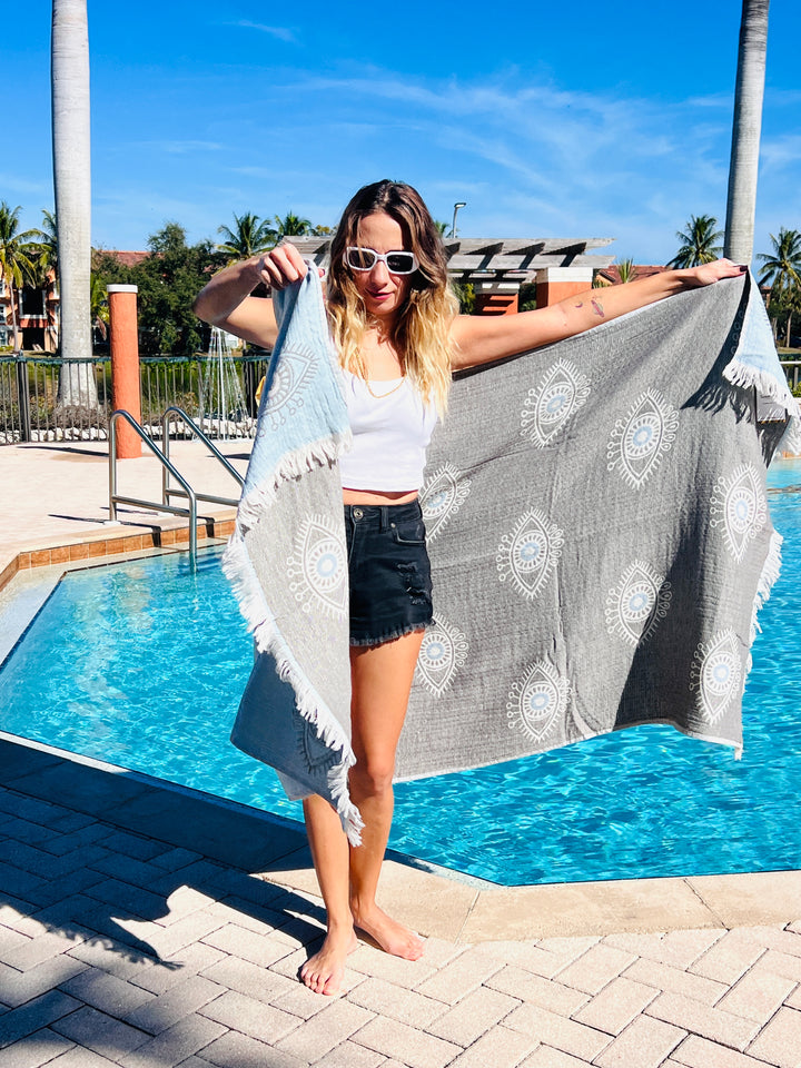 A woman stands barefoot by a pool, holding the Eye Sand Resistant Turkish Beach Towel from turkanhome.com, featuring a circular pattern and made from recycled fibers. She wears sunglasses, a white tank top, and black shorts. Palm trees and blue sky are behind her.
