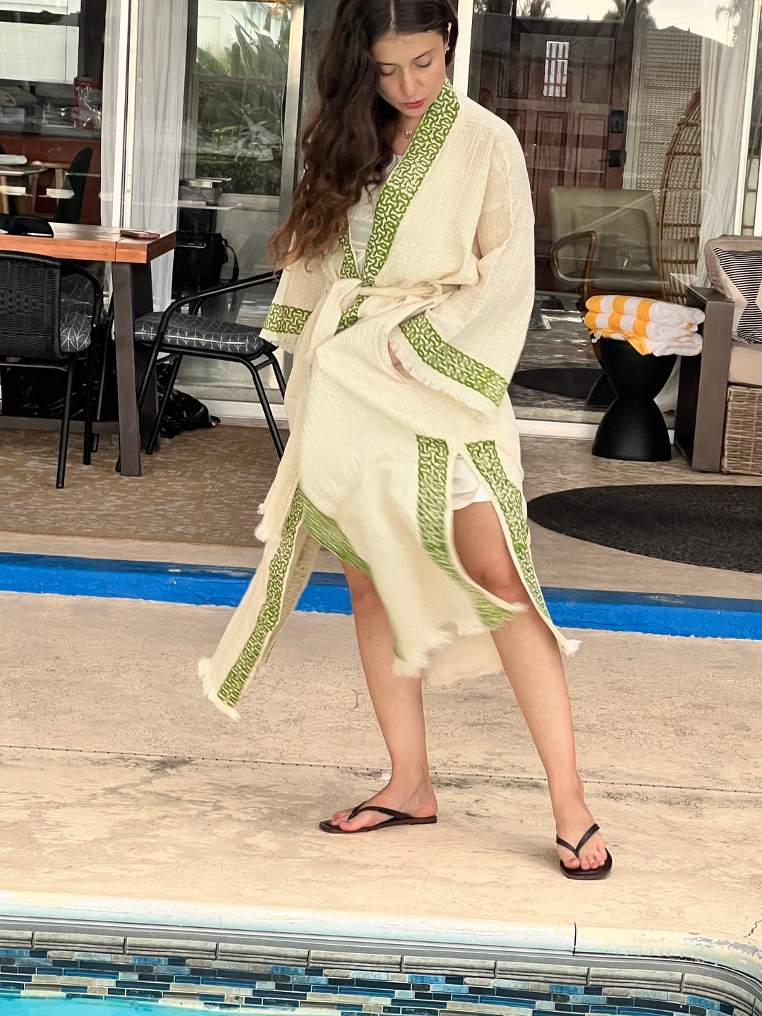 A woman stands poolside in flip-flops, wearing the Boho Kimono Robe – Poolside Beach Cover Up from turkanhome.com, featuring cream and green cotton muslin with frayed edges and tribal embroidery. Nearby are chairs, a table, and towels by a glass door.
