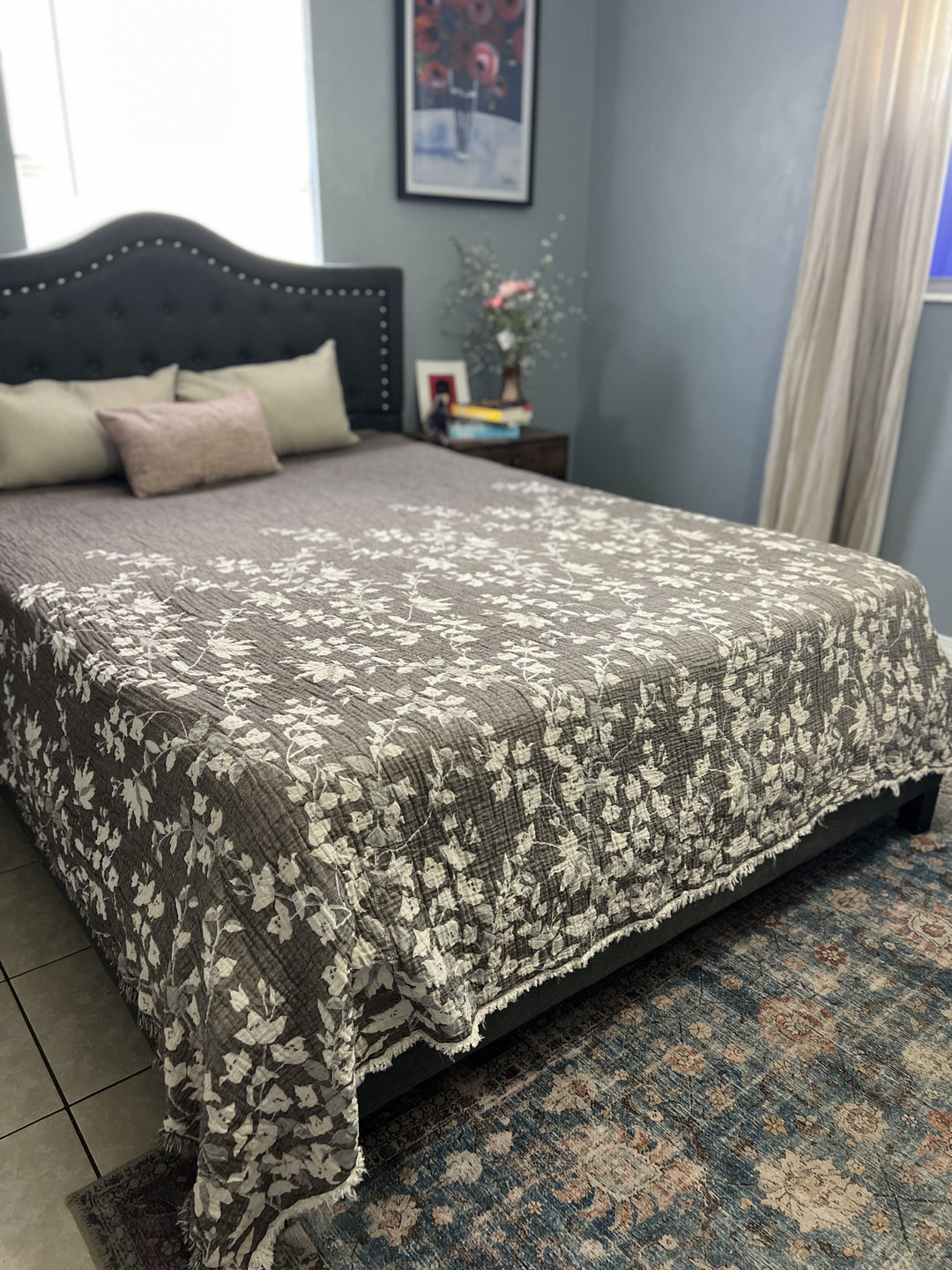 A neatly made bed with a gray and white floral bedspread, decorative pillows, and a dark tufted headboard features the Bougainvillea Muslin Bed Blanket in brown (Queen) from turkanhome.com. A nightstand with flowers, books, and a framed photo sits beside the bed.
