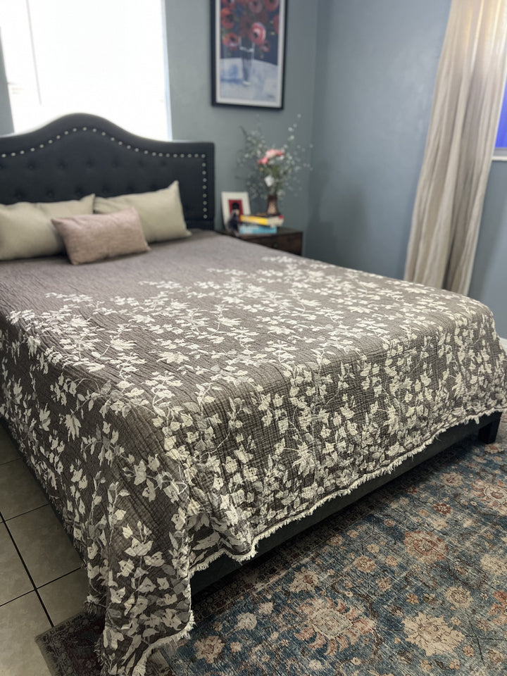 A neatly made bed with a gray and white floral bedspread, decorative pillows, and a dark tufted headboard features the Bougainvillea Muslin Bed Blanket in brown (Queen) from turkanhome.com. A nightstand with flowers, books, and a framed photo sits beside the bed.