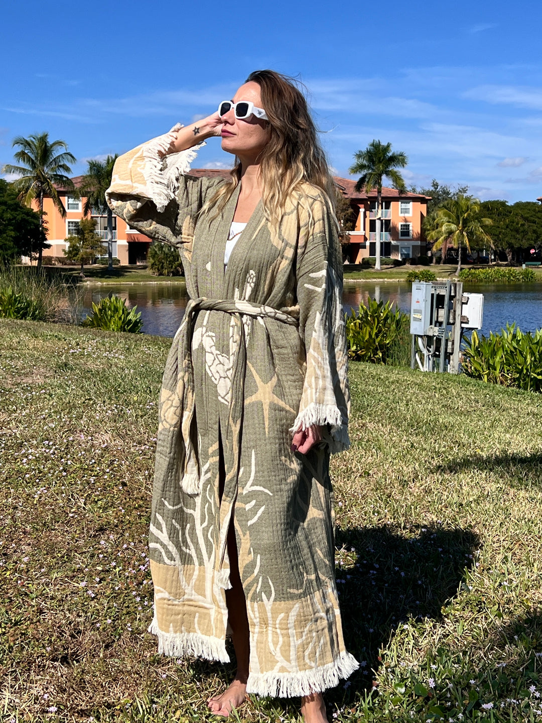 Wearing the Green Turtle Robe Kimono from turkanhome.com, a woman in white sunglasses stands on grass by a pond and palm trees, enjoying the sun in chic beachwear that radiates effortless boho loungewear style.