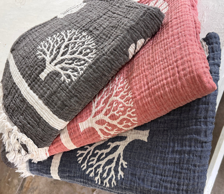A trio of folded Coastal Muslin Throw Blankets by turkanhome.com, each featuring a charming white tree design, brings cozy coastal vibes to any space. Crafted from soft, reversible Turkish cotton muslin, these stylish throws come in dark gray, pink, and navy blue—each color providing a striking backdrop for the tree motif. Perfect as an elegant beach blanket or for adding comfort at home.
