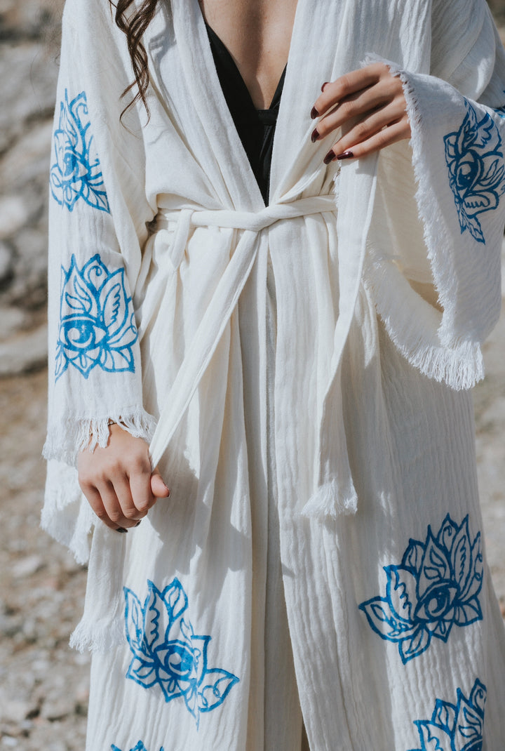 A woman wears the Lotus Kimono Robe by turkanhome.com—a white boho loungewear piece with blue floral patterns, frayed sleeves, and a fabric belt—styled over black clothing in an outdoor rocky setting.