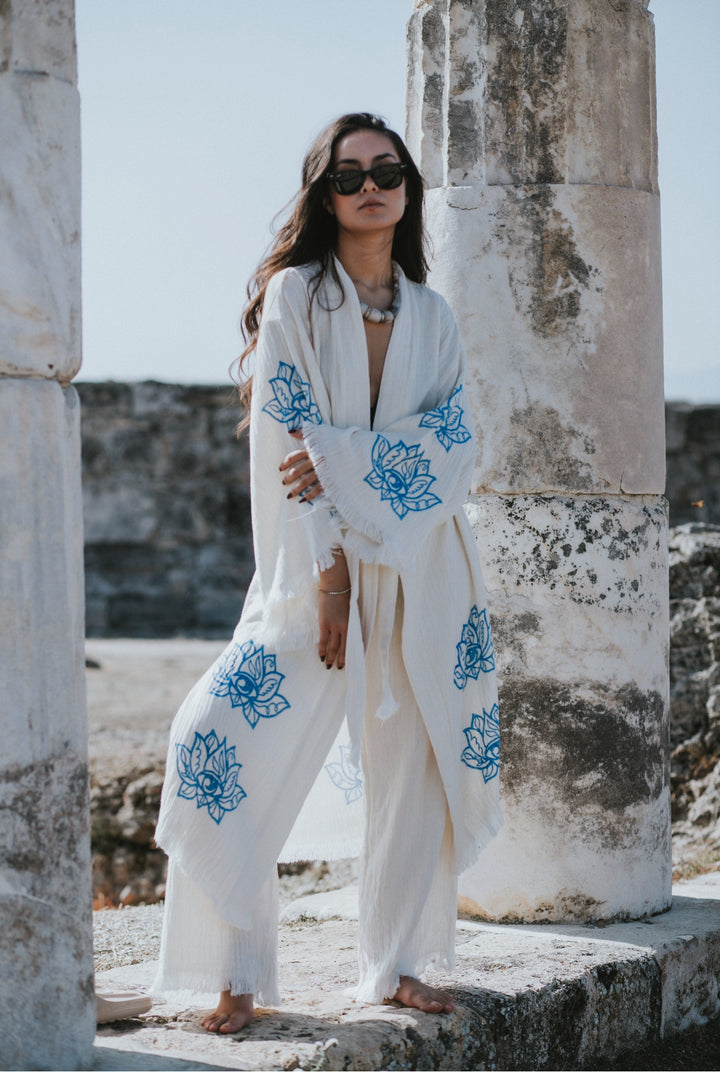 A woman in a Lotus Kimono Robe from turkanhome.com, featuring blue floral patterns, stands barefoot among ancient ruins, her sunglasses catching the sunlight as she highlights the eco-friendly elegance of this festival boho kimono.