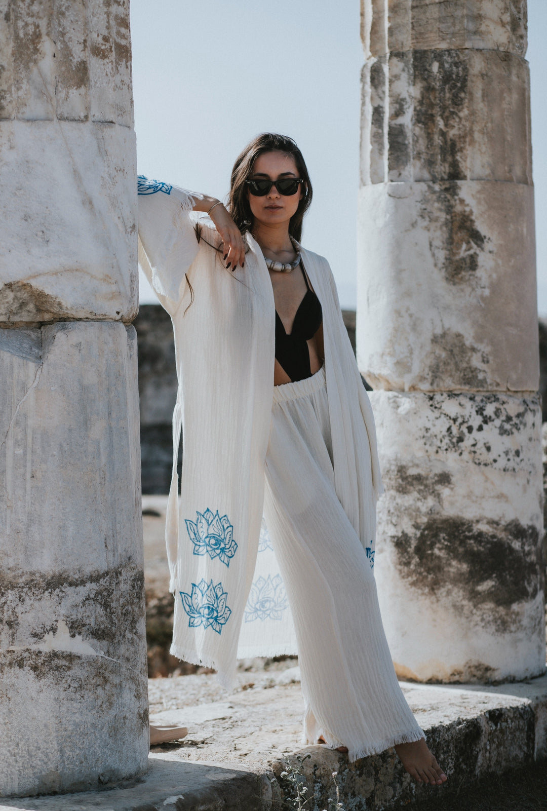A woman in sunglasses, a black top, and the Lotus Kimono Robe by turkanhome.com—featuring flowing white fabric with blue floral designs—leans against ancient stone columns beneath a bright sky.