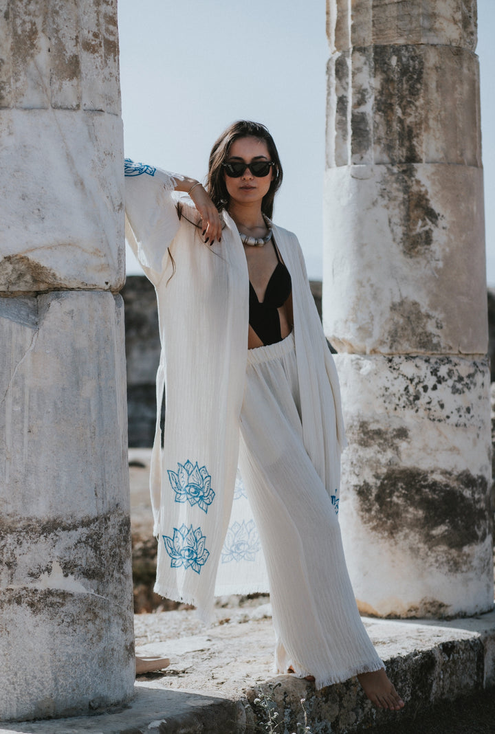 A woman in sunglasses, a black top, and the Lotus Kimono Robe by turkanhome.com—featuring flowing white fabric with blue floral designs—leans against ancient stone columns beneath a bright sky.