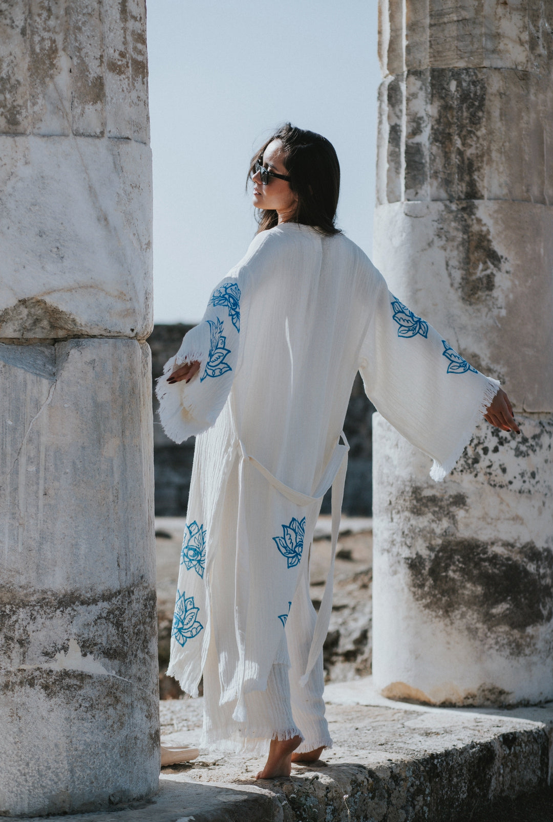 A woman in the Lotus Kimono Robe by turkanhome.com, featuring blue floral bamboo fiber fabric, stands barefoot between ancient stone columns. Sunlight highlights her eco-friendly loungewear and boho style as she looks back in sunglasses.