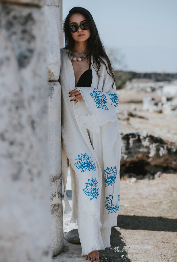 A woman in sunglasses and a Lotus Kimono Robe from turkanhome.com leans against a stone wall outdoors, surrounded by rocky terrain and a clear sky.