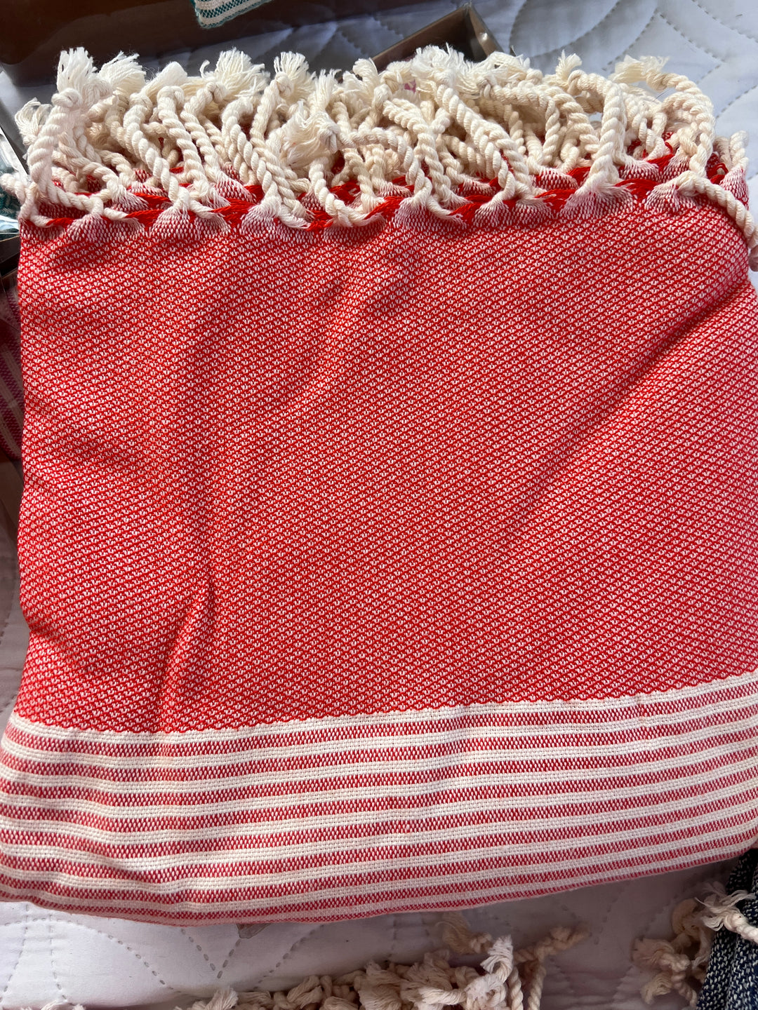 The Vegas Classy Bamboo Beach Towel from turkanhome.com features a folded red and white design with cream fringe, white stripes at the bottom, and a textured look. Its displayed on a quilted white surface.