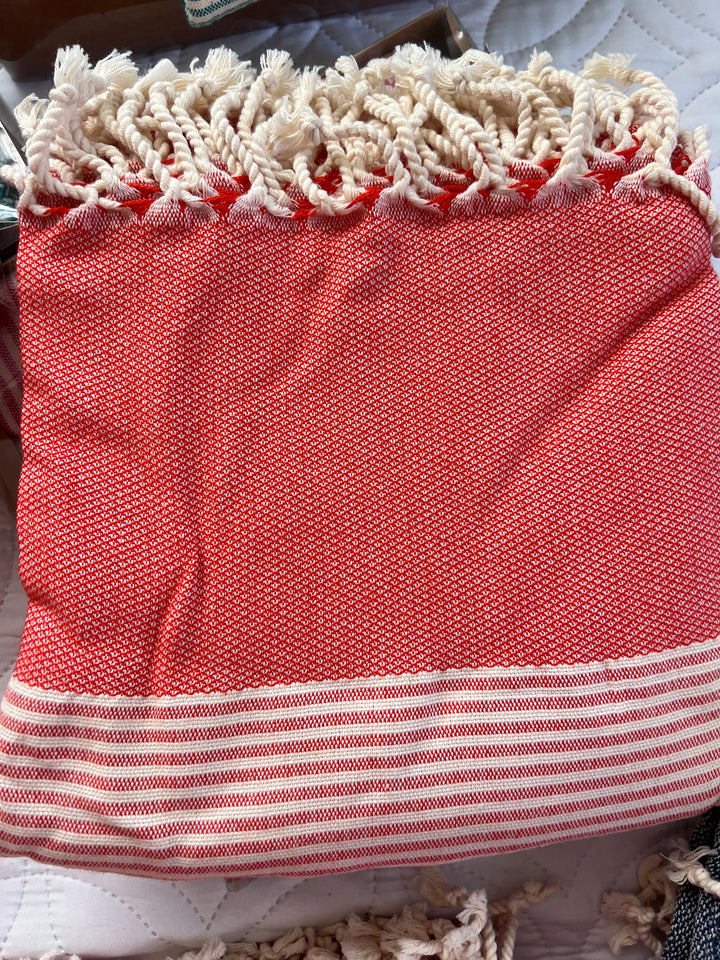 The Vegas Classy Bamboo Beach Towel from turkanhome.com features a folded red and white design with cream fringe, white stripes at the bottom, and a textured look. Its displayed on a quilted white surface.