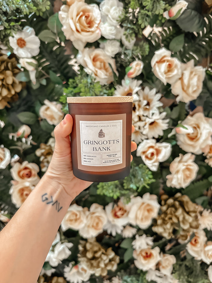 A hand with a GRYFF tattoo holds the Ampersand Candles + Oils Gringotts Bank Beeswax Candle (10oz) in front of cream roses and green foliage—an organic beeswax Harry Potter piece for any collection.