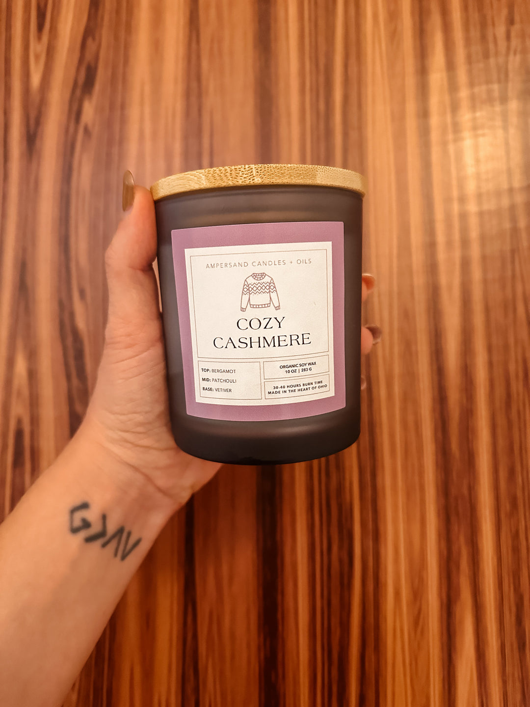 A tattooed hand holds a 10oz Cozy Cashmere Beeswax Candle by Ampersand Candles + Oils against a wood-patterned background.
