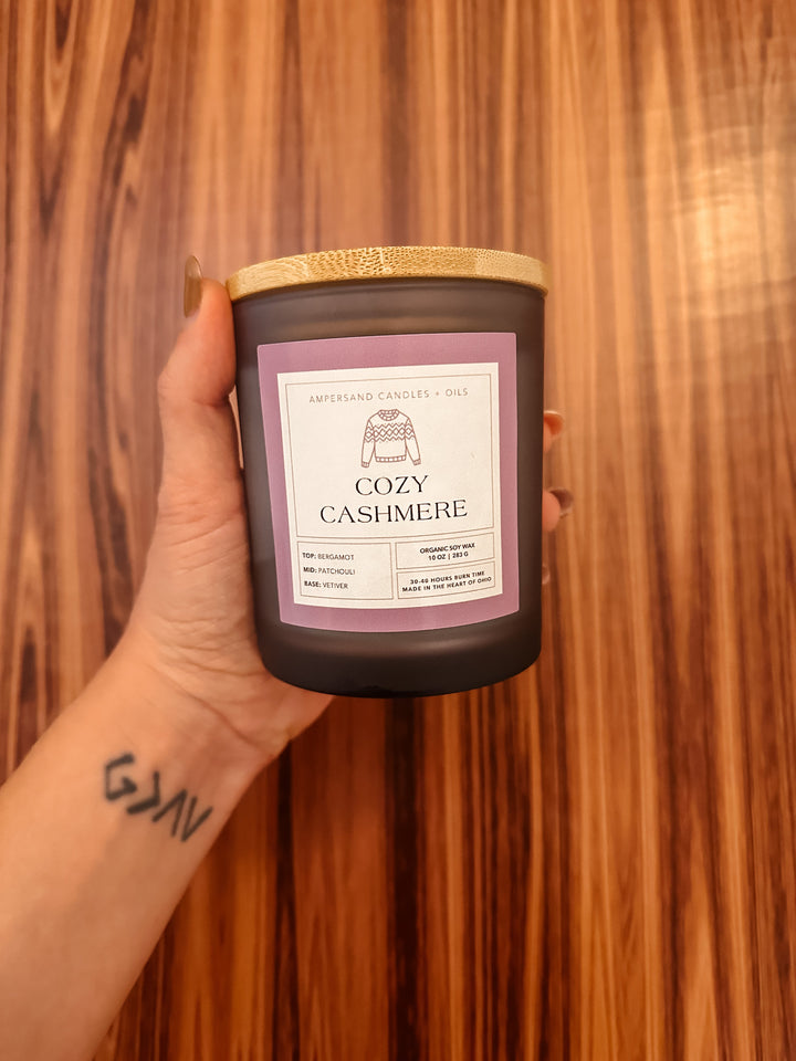 A tattooed hand holds a 10oz Cozy Cashmere Beeswax Candle by Ampersand Candles + Oils against a wood-patterned background.