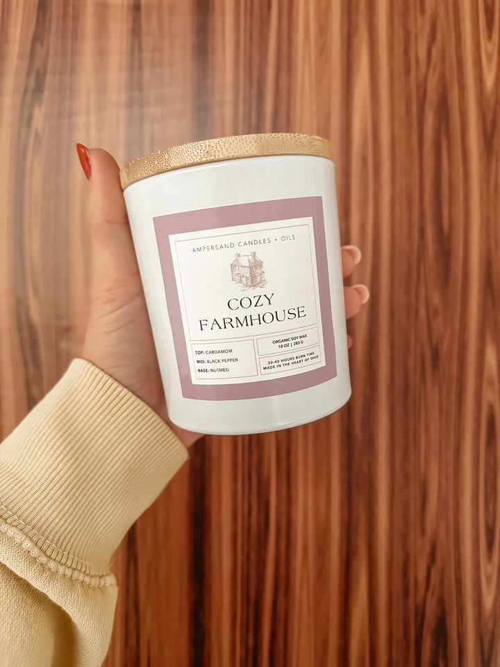 A hand with painted nails holds an Ampersand Candles + Oils Cozy Farmhouse Beeswax Candle (10oz) in a glass jar. The label highlights cardamom, black pepper, nutmeg, and vanilla notes in this organic candle against a wood-patterned backdrop.