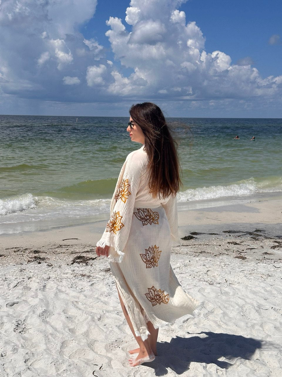 A woman with long hair stands barefoot on a sandy beach in the Lotus Kimono Robe from turkanhome.com, its white and gold eco-friendly fabric flowing as she faces the ocean beneath a partly cloudy sky.
