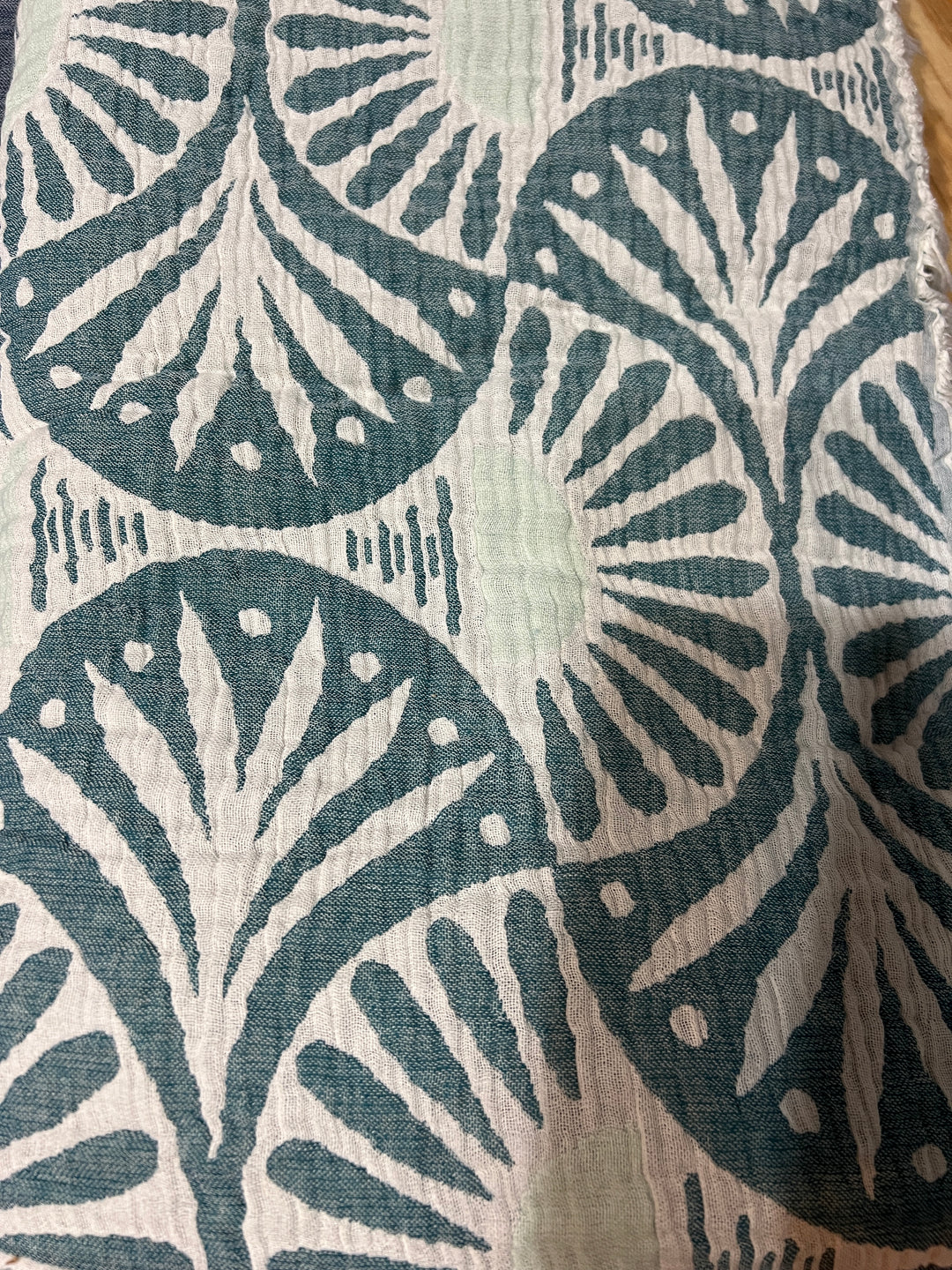 A close-up of the Gingko Muslin Bed Blanket – Teal (King) from turkanhome.com shows softly crinkled, breathable fabric with repeating teal and white fan and dot motifs, evoking the luxury of Turkish cotton.