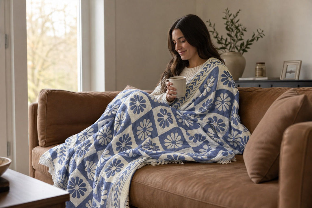A woman sits on a brown sofa, wrapped in the Daisy Muslin Throw Blanket from turkanhome.com, holding a mug and smiling. A potted plant and framed photo are on the side table in the background.