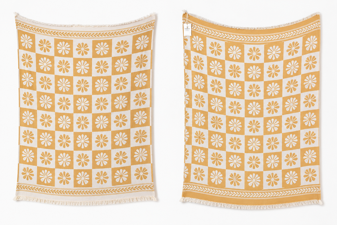 Two Daisy Muslin Throw Blankets from turkanhome.com in yellow and white hang side by side, each with a checkered pattern of floral motifs and decorative borders, displayed against a plain white background.