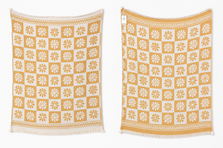 Two Daisy Muslin Throw Blankets from turkanhome.com in yellow and white hang side by side, each with a checkered pattern of floral motifs and decorative borders, displayed against a plain white background.