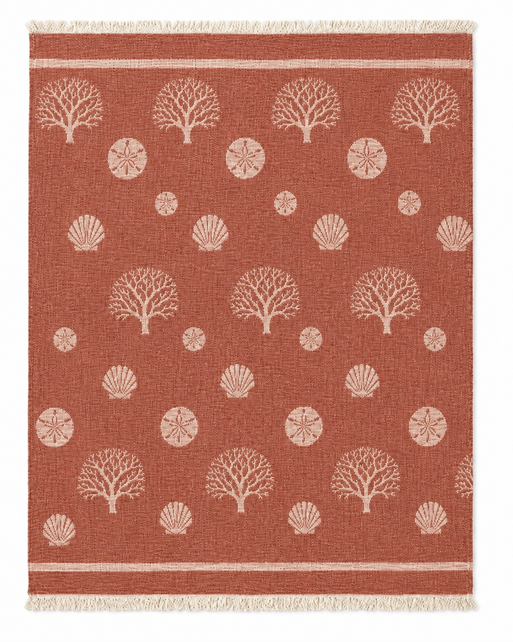 Wrap yourself in the Coastal Muslin Throw Blanket – Reversible Turkish Cotton from turkanhome.com, featuring a rectangular rust-colored design with light tan tree, shell, and sand dollar motifs, fringe on the short edges, and two parallel lines near each end—crafted from soft Turkish muslin cotton for a cozy beach blanket feel.