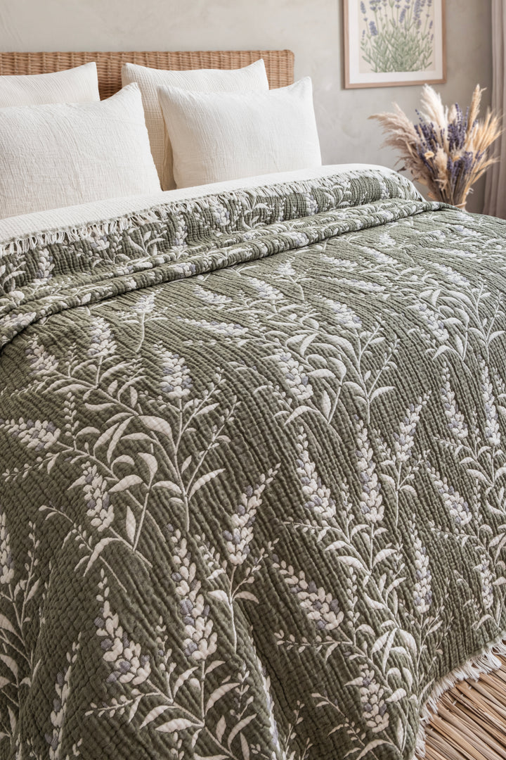 Olive green 7-Layer Cotton Muslin Quilted Blanket Lavander with a white floral pattern and fringe trim, draped over a bed with white pillows.