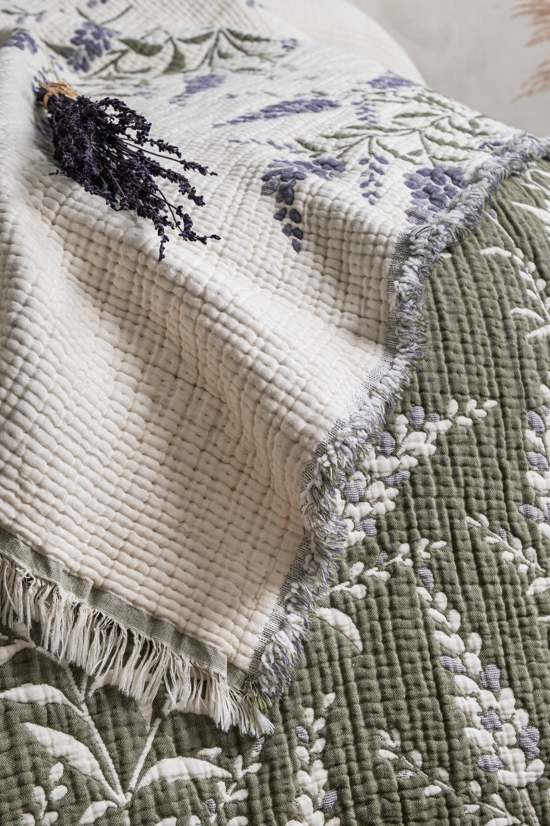A lavender 7-Layer Cotton Muslin Quilted Blanket Lavander with fringed edges, folded with a sprig of dried lavender on top, perfect as an all-season bedspread.