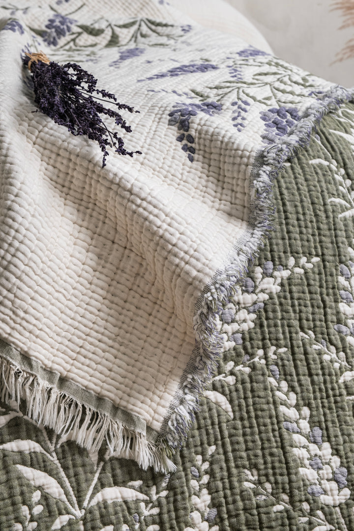 A lavender 7-Layer Cotton Muslin Quilted Blanket Lavander with fringed edges, folded with a sprig of dried lavender on top, perfect as an all-season bedspread.