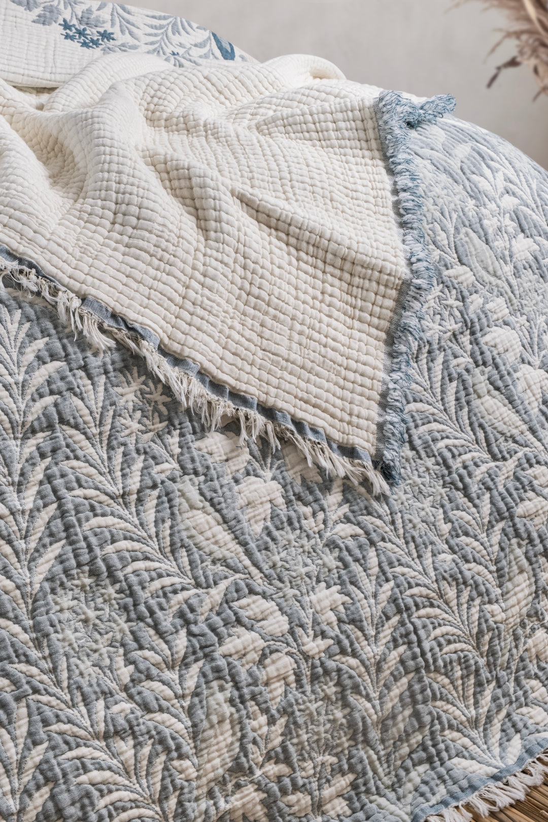 A blue and white leaf-patterned Luxury 7-Layer Cotton Muslin Quilted Blanket - Mint Bird comforter with a fringed edge folded over to reveal a plain cream-colored textured underside.