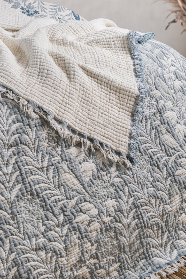 A blue and white leaf-patterned Luxury 7-Layer Cotton Muslin Quilted Blanket - Mint Bird comforter with a fringed edge folded over to reveal a plain cream-colored textured underside.