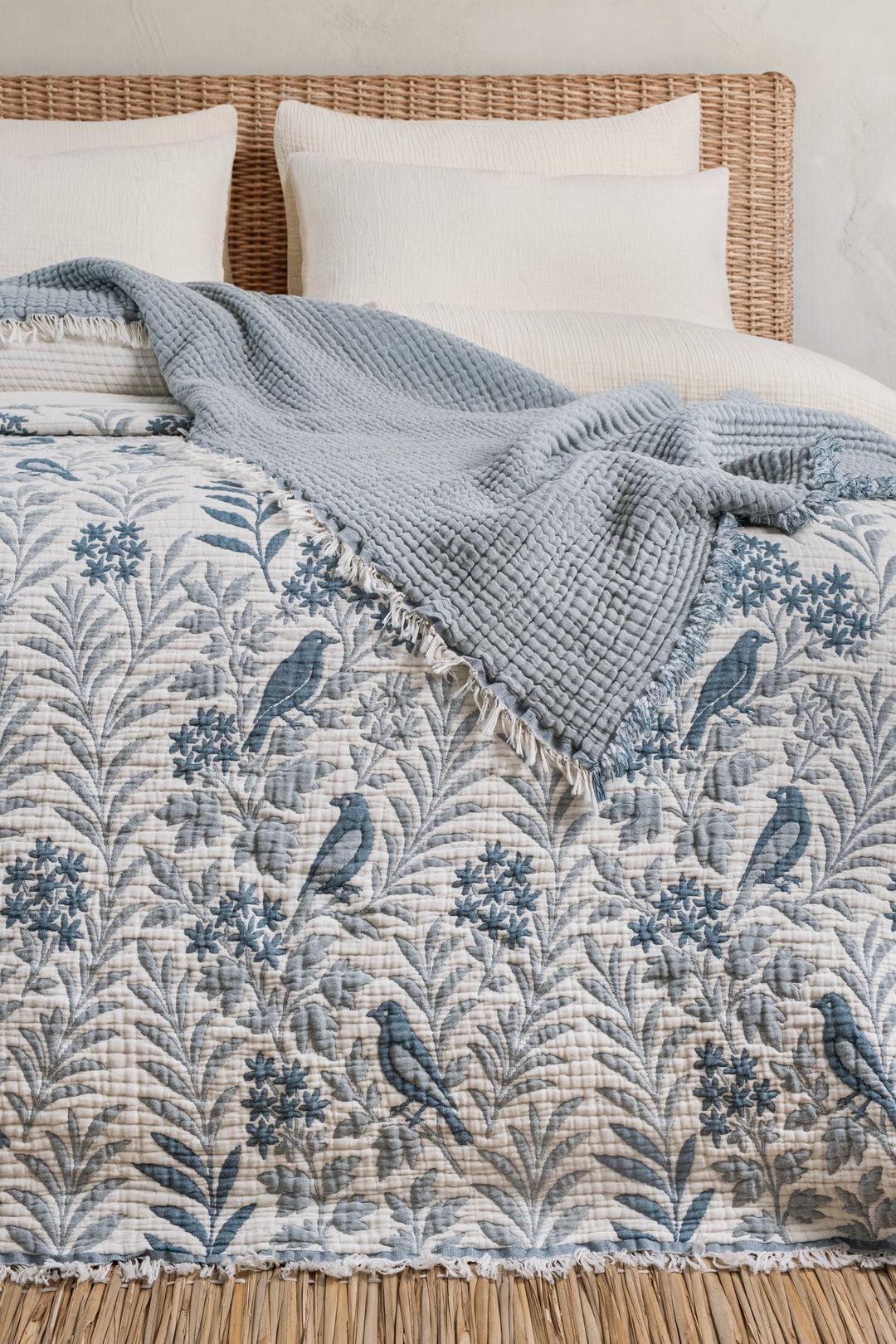 A Luxury 7-Layer Cotton Muslin Quilted Blanket - Mint Bird featuring a blue bird and floral pattern, partially covered by a solid light blue textured throw blanket on a bed.