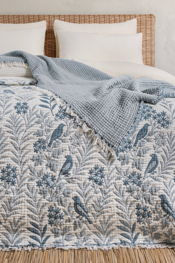 A Luxury 7-Layer Cotton Muslin Quilted Blanket - Mint Bird featuring a blue bird and floral pattern, partially covered by a solid light blue textured throw blanket on a bed.