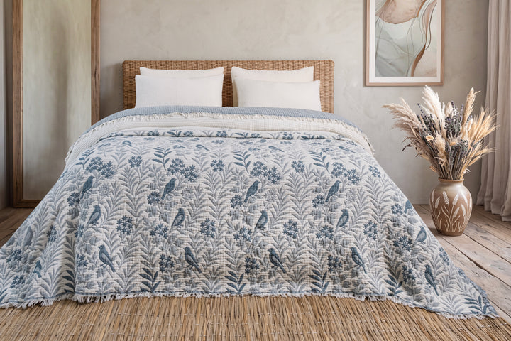 A Luxury 7-Layer Cotton Muslin Quilted Blanket - Mint Bird with a blue and white bird and floral print draped over a bed with white pillows and a woven headboard.
