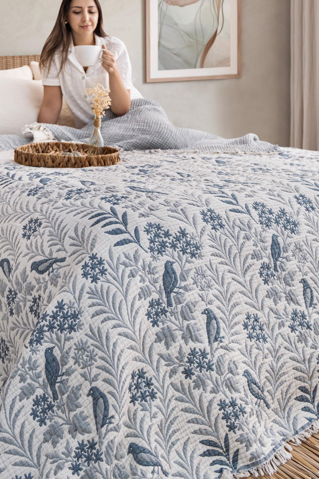 A white 7-layer Luxury 7-Layer Cotton Muslin Quilted Blanket - Mint Bird with a repeating blue print of birds and floral vines is draped over a bed where a woman sits drinking from a mug.