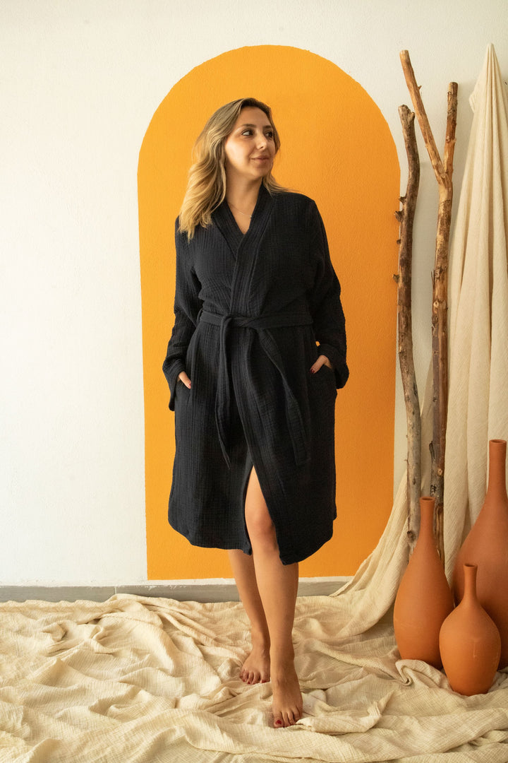 A woman with wavy blonde hair stands barefoot on a beige cloth, wearing the Cloud Muslin Robe by turkanhome.com. Behind her are an orange arch on the wall, wooden sticks, and terracotta vases. She glances to the side with a gentle smile.