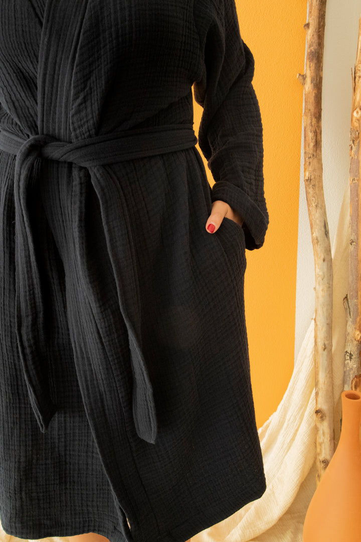 Wearing the Cloud Muslin Robe from turkanhome.com, a person stands against a yellow-orange backdrop with one hand in their pocket, showing red nails. A wooden stick and brown vase are nearby.