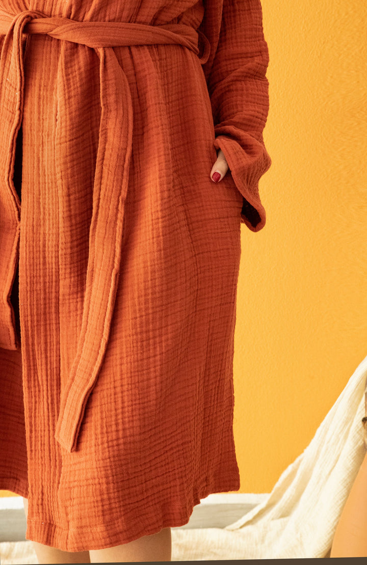 A person models the Cloud Muslin Robe by turkanhome.com, an orange Oeko-Tex certified bathrobe. Their torso and part of their legs are shown, one hand in a pocket, standing against an orange wall.