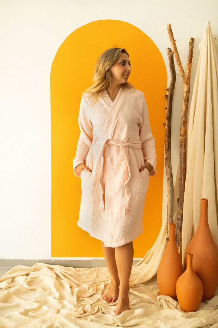 A woman wearing the Cloud Muslin Robe from turkanhome.com stands barefoot on a beige cloth, posed before a vibrant orange arch with tall wooden sticks and terracotta vases displayed to her right.