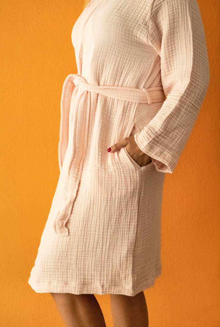 A person models the Cloud Muslin Robe by turkanhome.com in light pink with a textured finish and belted waist, standing against an orange wall—one hand in a pocket, face partially out of view.