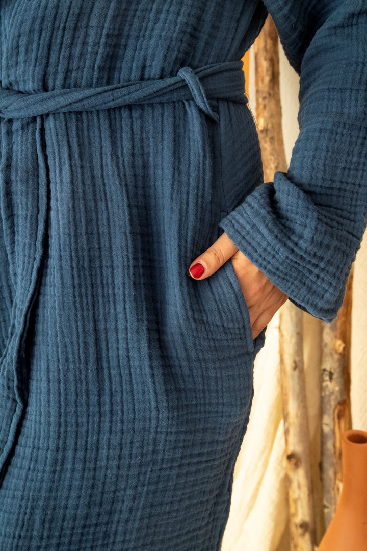 Close-up of a person in a blue Cloud Muslin Robe by turkanhome.com, Oeko-Tex certified with a belt and pockets. Red nail polish pops against the soft robe. Background is blurred wood and a clay vase.