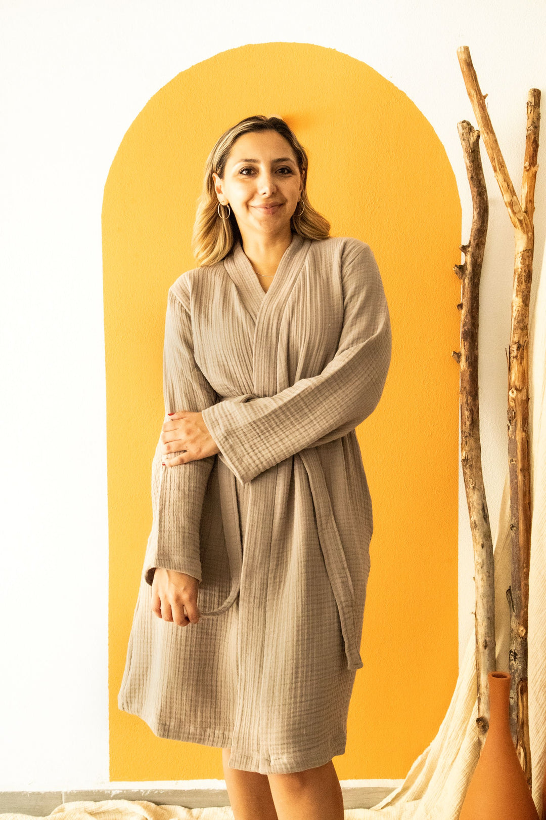 A woman, smiling with one arm crossed, stands before an orange arch wearing the Cloud Muslin Robe by turkanhome.com. Wooden sticks rest against the wall beside her.