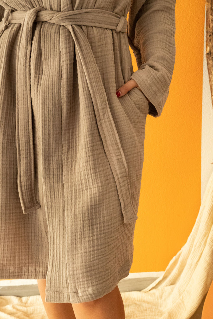A person models the Cloud Muslin Robe by turkanhome.com, showing its textured beige Turkish cotton fabric and belted design against an orange wall. Only the lower half of the robe and both hands—one in a pocket—are visible.