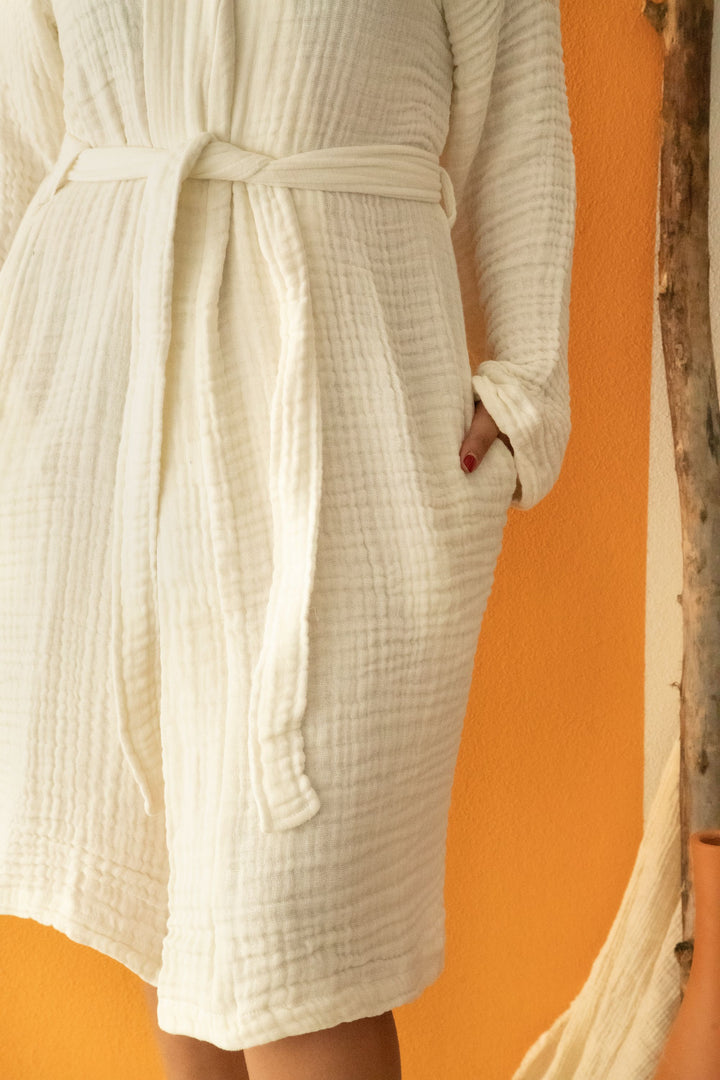 A person (face not shown) stands by an orange wall wearing the Cloud Muslin Robe from turkanhome.com—an Oeko-Tex certified, textured bathrobe with a belted waist and one hand in its pocket.