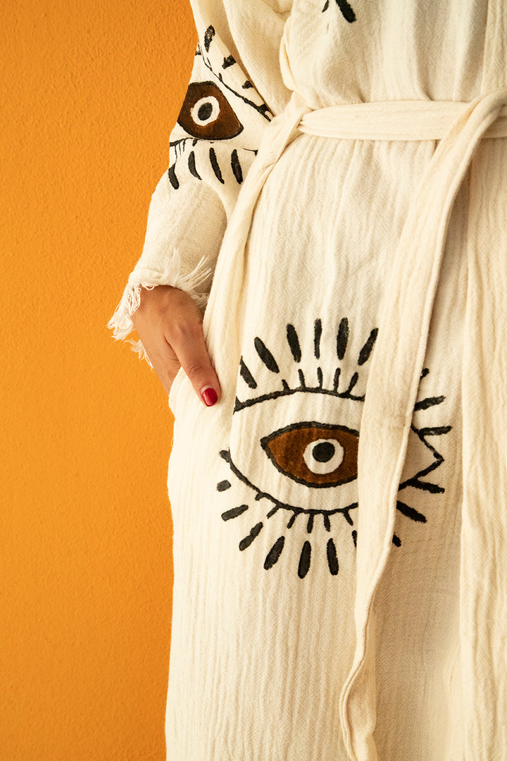 A person wearing the Brown Eye Kimono Robe from turkanhome.com, featuring eco-friendly cream fabric with bold black and brown eye motifs, rests a hand in the pocket against a solid orange wall.