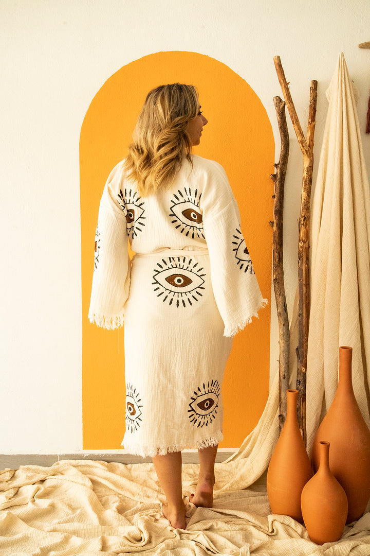 A woman stands barefoot on a textured rug, facing away, wearing the Brown Eye Kimono Robe by turkanhome.com—cream cotton with black eye motifs and fringe. An orange arch, wooden sticks, and clay vases are nearby.