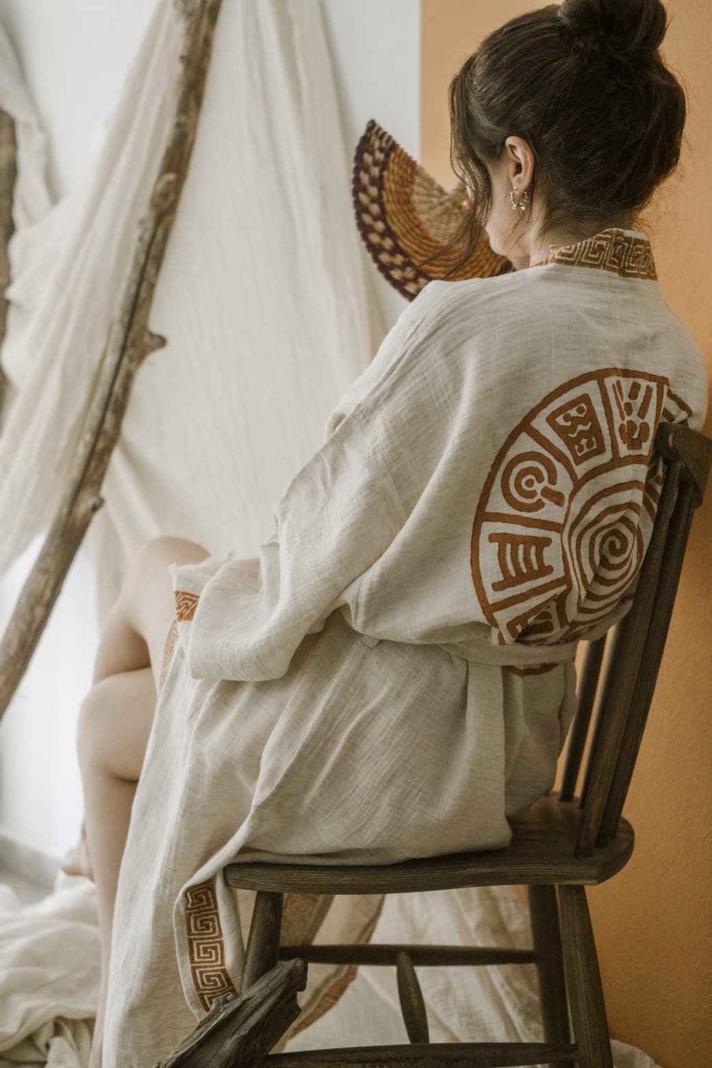 A woman with brown hair in a bun sits sideways on a wooden chair near white drapes and an orange wall, wearing the Triable Boho Kimono Robe - Poolside Beach Cover Up from turkanhome.com, featuring tribal hand block print.